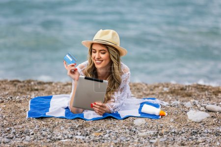 Téléchargez les photos : Jeune fille commander quelque chose en ligne whith la carte, tout en étant couché sur la plage - en image libre de droit