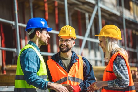 Foto de Un grupo de tres trabajadores en un sitio de construcción con sombreros duros, gafas de seguridad y ropa reflectante, sonriendo y conversando. - Imagen libre de derechos
