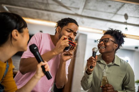 Foto de Grupo multirracial de amigos teniendo una noche de karaoke. Cantar en el micrófono, beber cerveza y tocar el órgano de la boca. - Imagen libre de derechos