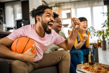 Téléchargez les photos : Groupe multiracial d'amis regardant le match de basket, mangeant de la pizza, buvant de la bière et acclamant. - en image libre de droit