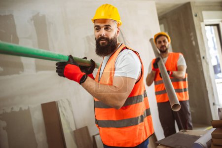 Téléchargez les photos : Deux travailleurs de la construction transportant des tuyaux sur un chantier - concepts de personnes au travail - en image libre de droit