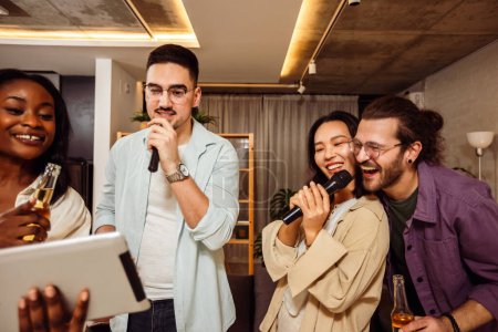 Foto de Grupo multirracial de amigos teniendo una noche de karaoke. Cantando en el micrófono, mirando letras y bebiendo cerveza. - Imagen libre de derechos