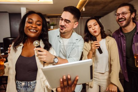 Téléchargez les photos : Groupe multiracial d'amis ayant nuit karaoké. Chanter au micro, regarder les paroles et boire de la bière. - en image libre de droit