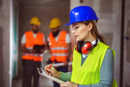 Photo for Portrait of successful woman constructor wearing yellow helmet and safety yellow vest. Portrait of architect standing at building site and writing something down on paper. - Royalty Free Image