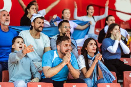 Foto de Los aficionados al deporte animando en el partido en el estadio. Usando colores azul y blanco para apoyar a su equipo. Celebrando con banderas y bufandas. - Imagen libre de derechos
