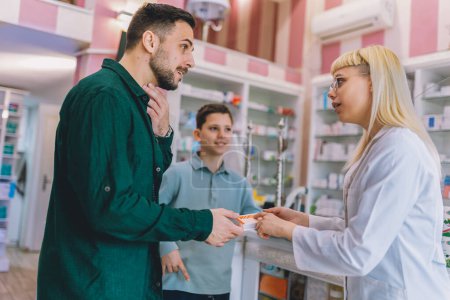 Photo for Cheerful young blonde pharmacist chemist woman giving vitamins, medicine to father and son. Having sore throat. - Royalty Free Image