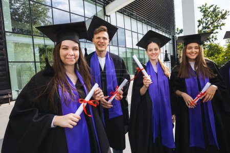 Foto de Grupo de graduados felices de pie frente a la escuela con togas y diplomas. - Imagen libre de derechos