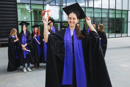 Foto de Retrato de estudiante graduada de pie frente a sus amigos con toga y diploma todo feliz. - Imagen libre de derechos