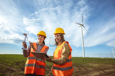 Photo for Two women engineers working and looking at the report wind turbine farm. They are wearing florescent vests and helmets for protection. - Royalty Free Image