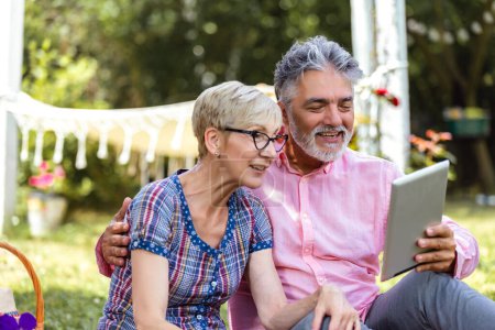 Porträt eines liebenden Senioren-Paares, das beim Picknick auf einer Decke sitzt und ein digitales Tablet benutzt