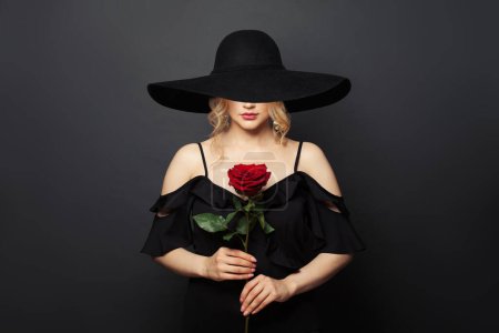 Foto de Hermosa mujer apasionada con amplio sombrero de ala ancha negro con flor de rosa roja sobre fondo negro - Imagen libre de derechos