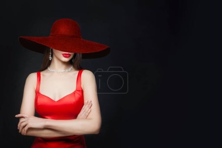 Foto de Joven morena con labios rojos maquillaje y collar de diamantes y pendientes y en sombrero ancho de color burdeos, retrato sobre fondo negro - Imagen libre de derechos