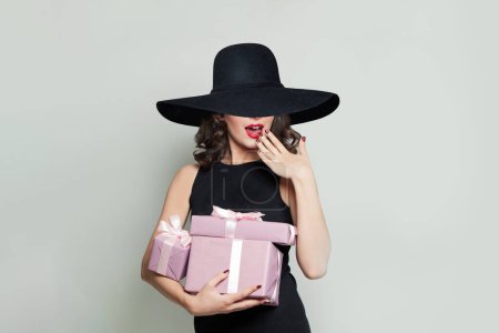 Foto de Mujer feliz sorprendida con regalos rosados y en negro wude sombrero de ala ancha sobre fondo gris claro - Imagen libre de derechos