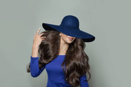 Foto de Joven mujer morena hermosa en vestido azul y amplio sombrero de ala ancha posando sobre fondo blanco - Imagen libre de derechos