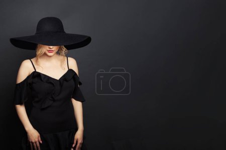 Foto de Mujer elegante con vestido negro y sombrero ancho de ala ancha negro sobre fondo negro - Imagen libre de derechos