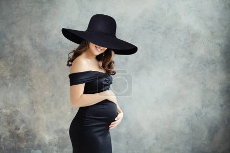 Foto de Retrato de belleza de moda de mujer embarazada hermosa en vestido negro y sombrero ancho negro sobre fondo gris - Imagen libre de derechos