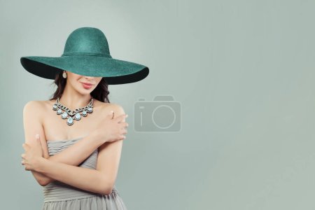 Foto de Hermosa mujer joven modelo de moda en vestido sedoso y amplio sombrero de ala ancha verde sobre fondo gris, retrato de moda - Imagen libre de derechos