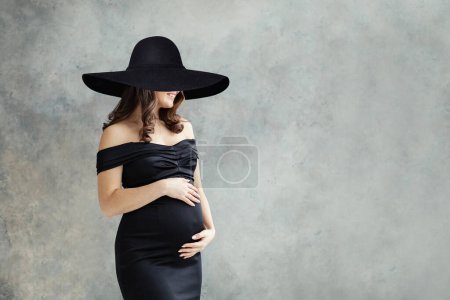 Foto de Mujer embarazada alegre en amplio retrato de sombrero de ala ancha negro. Hermosa dama morena vistiendo vestido negro anuncio sonriendo sobre fondo gris estudio - Imagen libre de derechos