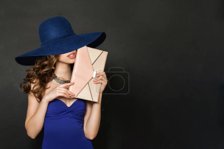 Foto de Hermosa mujer vestida con un guante de noche azul y un sombrero azul ancho. Mujer con pelo castaño, bolso de embrague y collar de joyería - Imagen libre de derechos