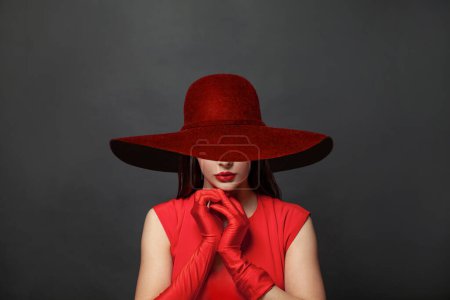 Foto de Mujer elegante morena con maquillaje y cabello oscuro con vestido rojo y guantes de seda roja y sombrero rojo de ala ancha sobre fondo negro - Imagen libre de derechos