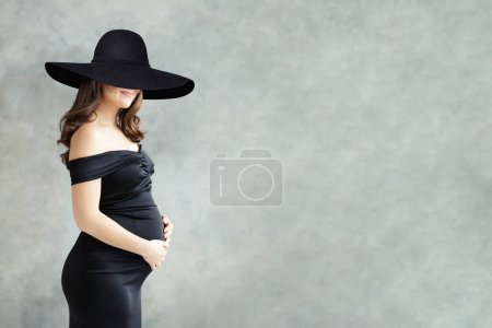 Foto de Retrato de moda de mujer embarazada en vestido negro y en amplio sombrero de ala ancha negro sobre fondo gris - Imagen libre de derechos