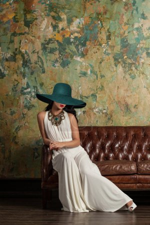 Foto de Hermosa modelo joven en vestido de noche blanco y sombrero de ala ancha verde sentado en un sofá de lujo - Imagen libre de derechos