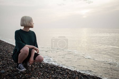 Téléchargez les photos : Jolie jeune femme adulte avec de courts cheveux blonds assis sur la plage de la mer en plein air - en image libre de droit