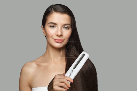 Téléchargez les photos : Brunette modèle femme tenant fer à repasser et redresser ses cheveux longs et foncés en bonne santé sur fond blanc. Coiffure et coiffure concept - en image libre de droit