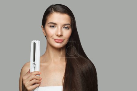 Beautiful model woman holding hair iron and straightening her healthy long dark hair on white background. Haircare and hair styling concept