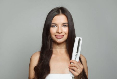 Téléchargez les photos : Nice young healthy brunette woman holding hair iron and straightening her long smooth shiny hair on white background. Haircare, hairstyle, hairdressing and hair styling concept - en image libre de droit