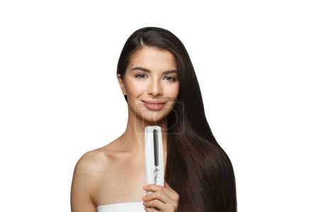 Téléchargez les photos : Happy young healthy brunette woman holding hair iron and straightening her long smooth shiny hair isolated on white background. Haircare, hairstyle, hairdressing and hair styling concept - en image libre de droit