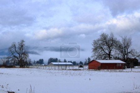 Die Landschaft eines Bauernhofs in der kalten, dunklen Wintersaison mit Wirtschaftsgebäuden, Schnee und bewölktem Himmel. 