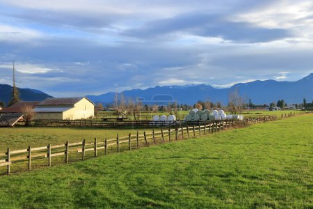 A long handmade wooden fence and a rural landscape with acres of grassland, farm buildings and hay bales.  