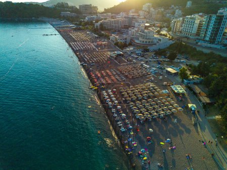 Photo for Wide sandy beach with sun umbrellas, plastic white sunbeds and people relaxing in swimsuits in the historic tourist resort town of Becici near Adriatic Sea against the backdrop of evening sunny sky - Royalty Free Image