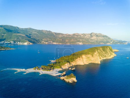 Historical ancient tourist island of St. Nicholas with vegetation on rocky shores in the azure Adriatic Sea against the backdrop of coastal resort towns, the Montenegrin mountain range and clear sky