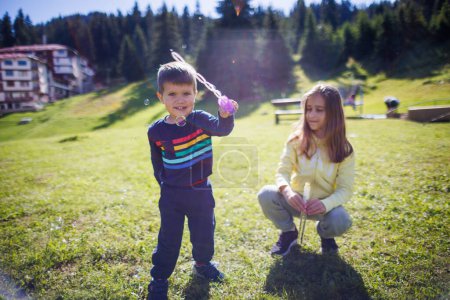 Téléchargez les photos : An older sister with long hair in a yellow tracksuit plays with her younger brother in a blue suit and teaches him how to blow soap bubbles on a green lawn in the backyard in spring - en image libre de droit