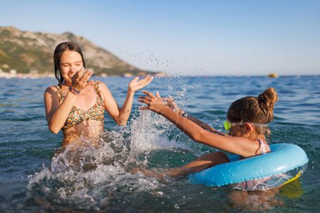 Foto de An older sister and her younger sister with blue swimming goggles, who sits in a small bright inflatable circle, splash each other in the warm waters of the Adriatic Sea in the evening sunlight - Imagen libre de derechos