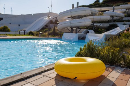 Téléchargez les photos : A yellow huge inflatable circle lies at the edge of a large pool with transparent blue shiny water against the background of a summer vegetable water park and a summer cloudy sunny sky - en image libre de droit