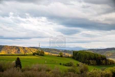 Photo for Tall telecommunications tower for communication in a hilly countryside with tall green grass and dry trees, against the backdrop of dark mountains and a cloudy gray sky in the evening sunlight - Royalty Free Image