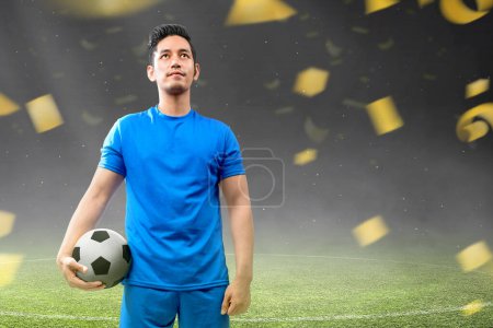 Foto de Un jugador de fútbol asiático en un jersey azul está de pie y sosteniendo la pelota en el campo de fútbol - Imagen libre de derechos