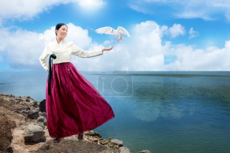 Téléchargez les photos : Femme asiatique portant un costume national coréen traditionnel, Hanbok, debout avec les paumes ouvertes et montrant oiseau volant au bord du lac avec fond bleu ciel - en image libre de droit