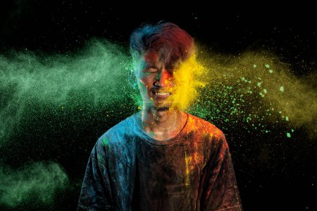 Photo for Asian man splashed with colorful holi powder and having fun isolated on black background. Color festival India concept. - Royalty Free Image
