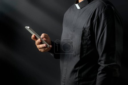 Téléchargez les photos : Portrait d'un pasteur utilisant un téléphone portable sur fond noir. Concept chrétien - en image libre de droit