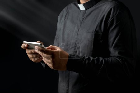 Téléchargez les photos : Jeune prêtre tenant le téléphone, textos quelque chose isolé sur fond noir. - en image libre de droit