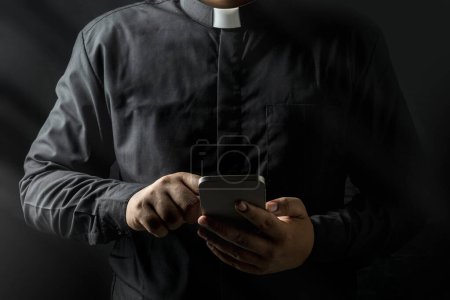 Photo for Young priest holding smartphone and touch the screen isolated on black background. - Royalty Free Image