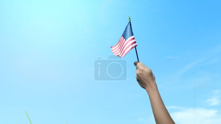 Foto de Closeup view of a human hand holding an American flag with a blue sky background. 4th of July concept - Imagen libre de derechos