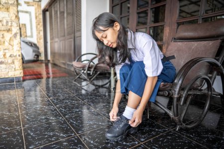 Photo for Young female Indonesian with junior hich school uniform going to school in the morning in front of the house tying shoelaces. Back to school concept. - Royalty Free Image