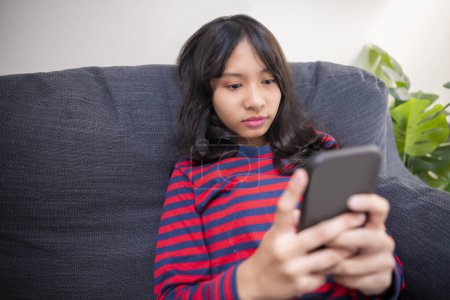 Foto de Asian girl sitting on the couch while using a mobile phone at home - Imagen libre de derechos