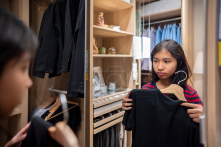 Foto de Indonesian pretty teenage girls choosing and thinking clothes while looking in the mirror in a closet at home. Asian female deciding what shirt to wear in the morning. Fashion shopping store concept. - Imagen libre de derechos