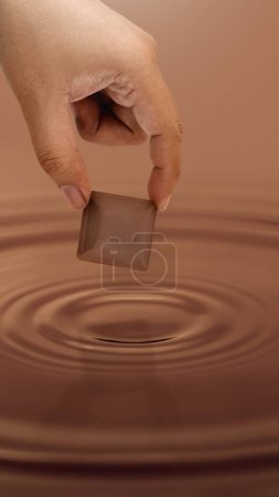 Foto de Close up human hand holding chocolate bar above the melted chocolate liquid, concept of giving on world chocolate day. Sweet and energy snack. Mock up deisign. - Imagen libre de derechos